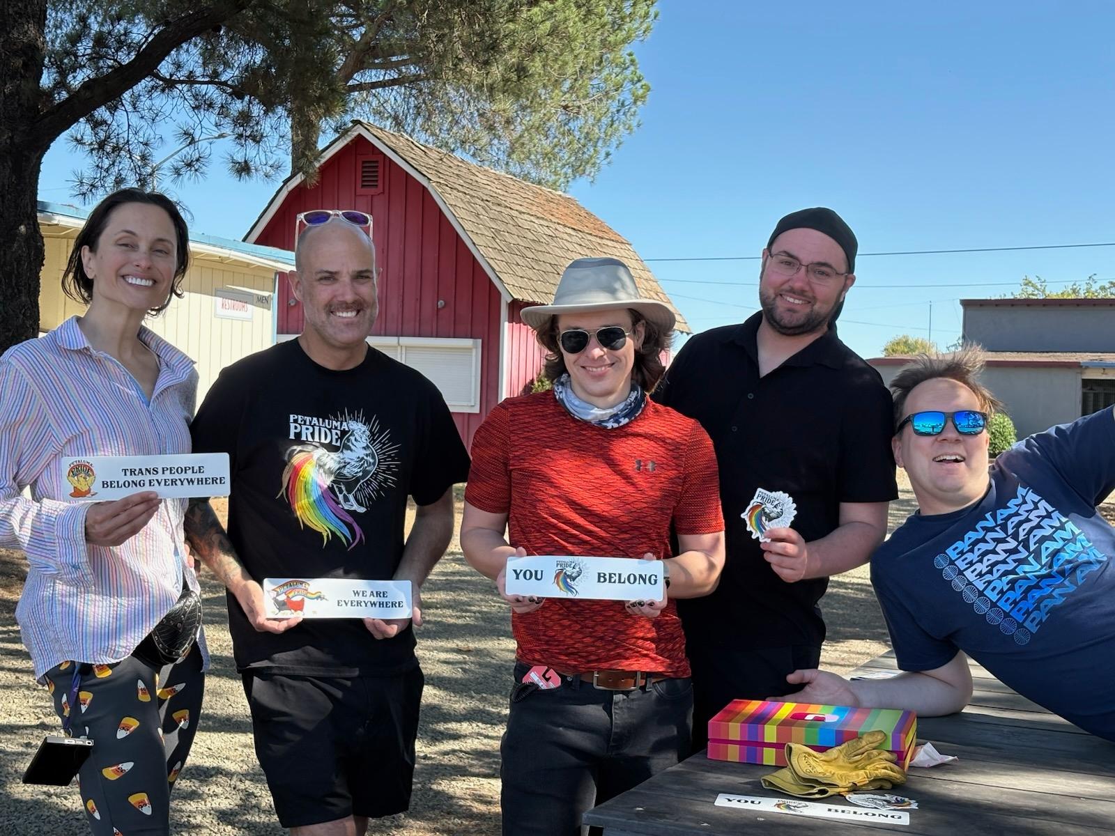 A photo of Jennifer, Eric, Robin, Andy, and Chris posing together in front of a red barn-like building at the fairgrounds under the shade of trees and a clear blue sky. Each is holding a pride bumper sticker including:
one with an illustration of a chick hatching and holding a Petaluma Pride banner, adjacent to text that reads: trans people belong everywhere 
another with an illustrated family of hens and chicks running with the Petaluma Pride banner, adjacent text that reads: we are everywhere 
another which the iconic Petaluma Pride illustrated rooster with rainbow plumage, flanked by the words: you belong 
and another that is just the Petaluma Pride rooster