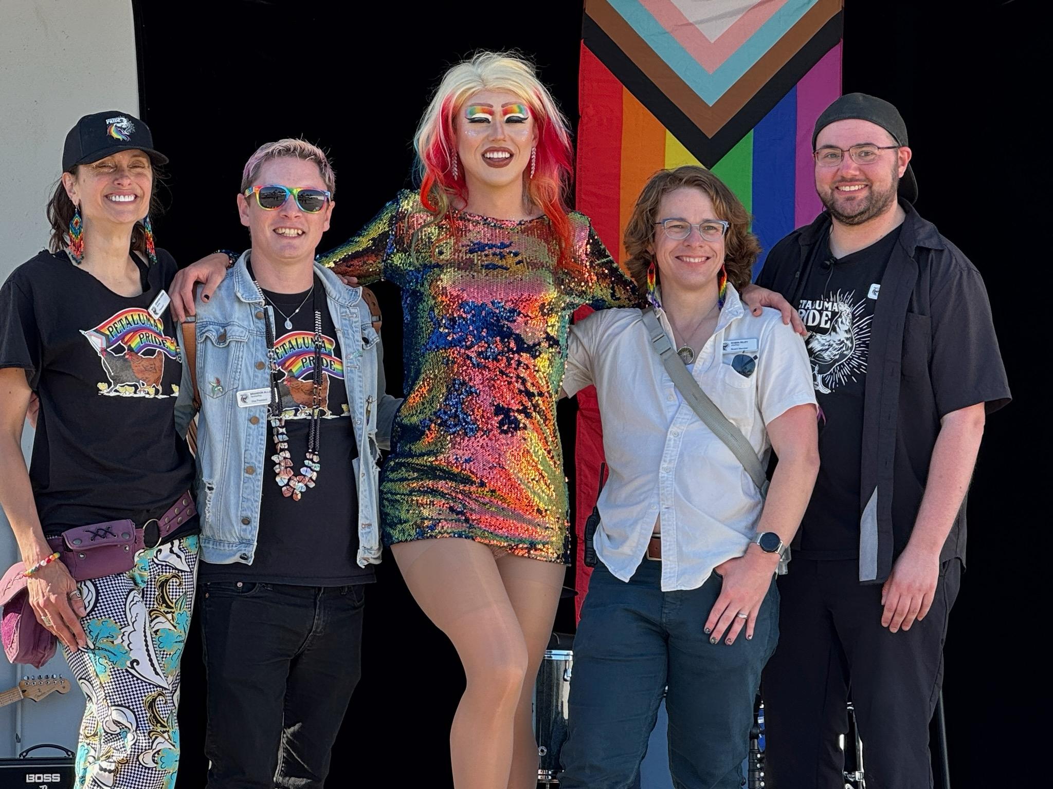 A photo of Jennifer, Brandon, Adda, Robin, and Andy with their arms around each other in front of a stage backdrop featuring a giant progress pride flag. Everyone is smiling and dressed in pride gear.