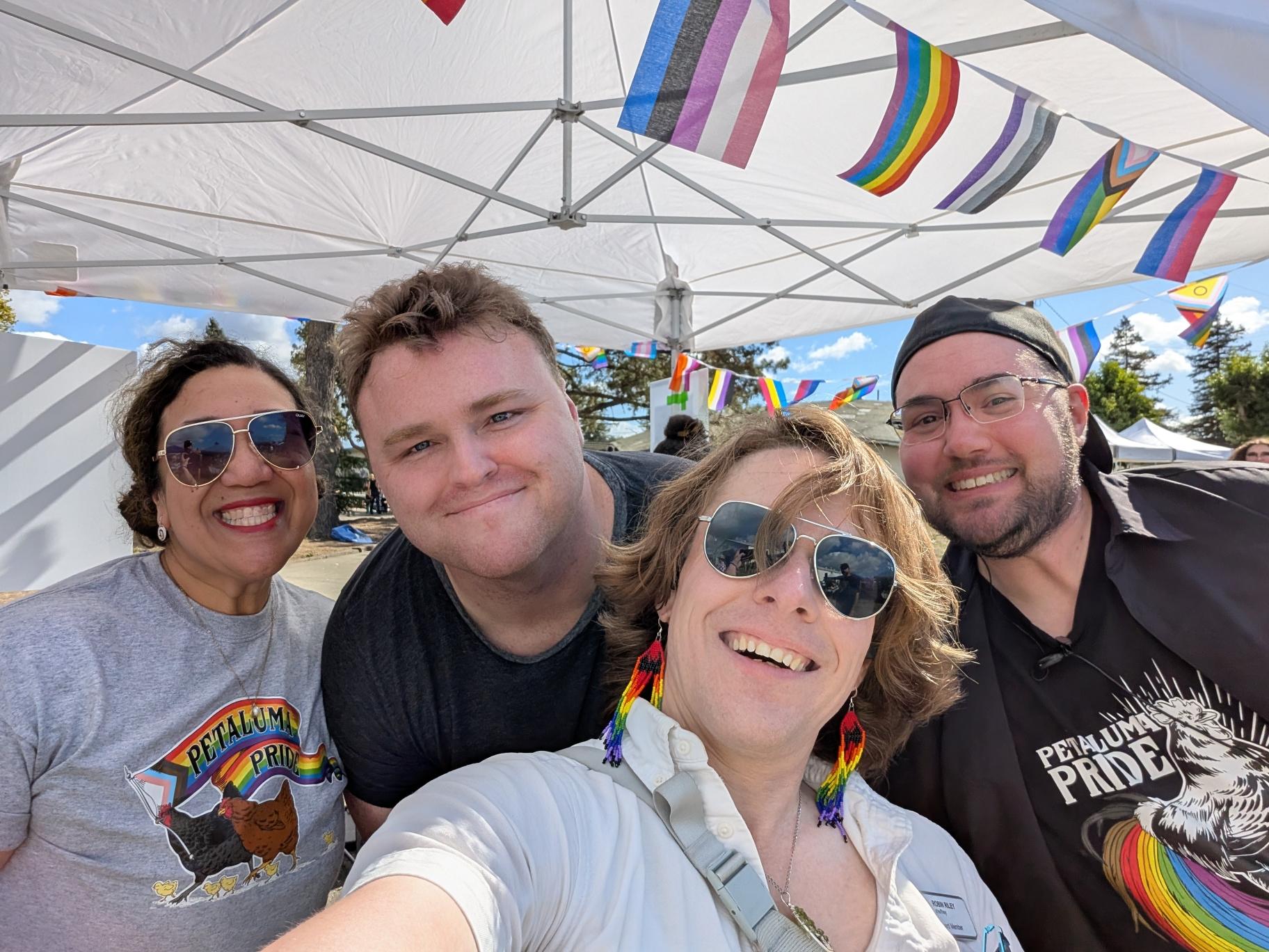 A group selfie of Mo, Blake, Robin, and Andy under a pop-up canopy on a sunny afternoon. Everything about the photo is extremely colorful, as is appropriate for Pride! Robin's wearing rainbow beadwork dangly earrings featuring bats.