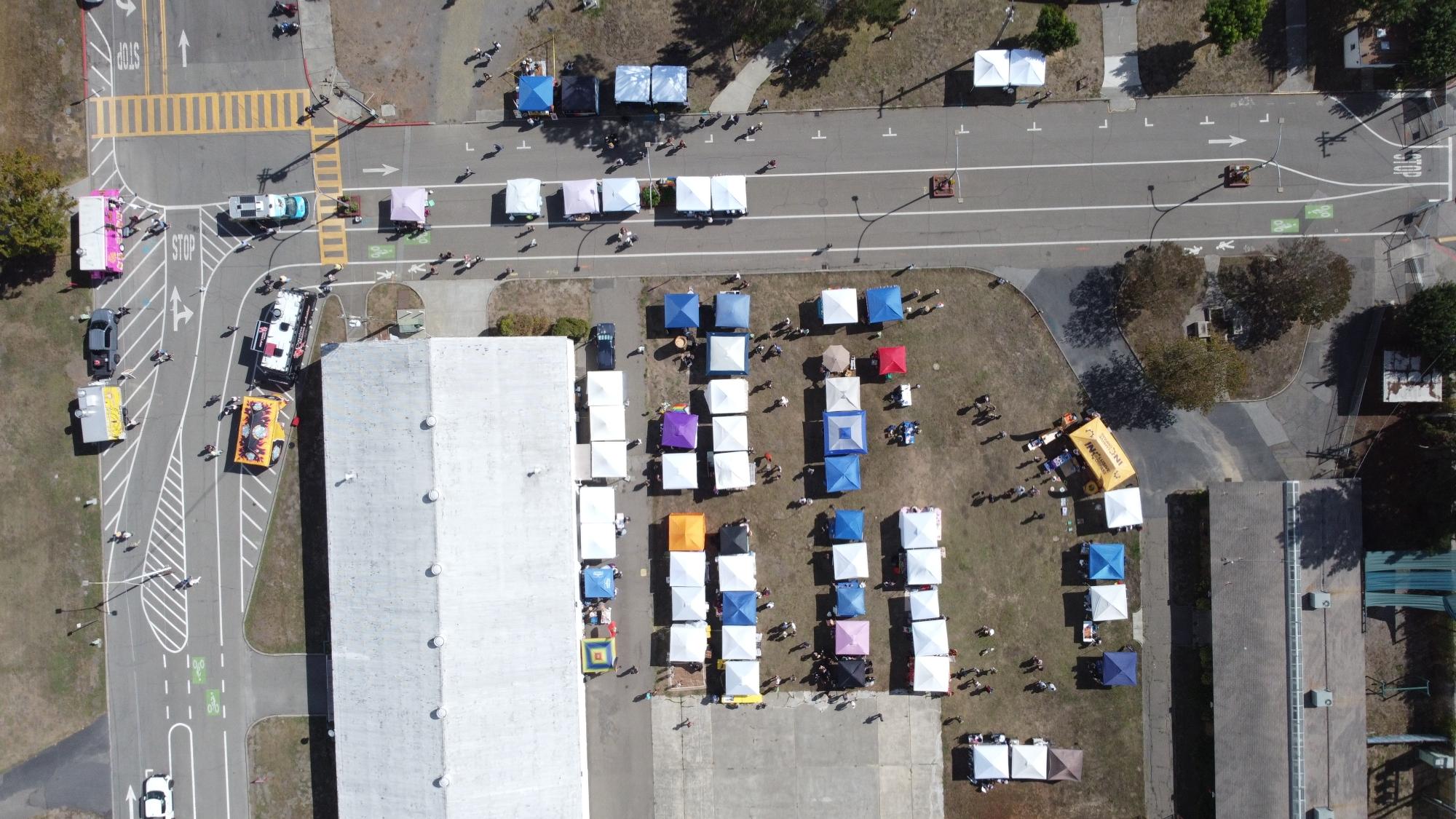 An aerial photo of much of the pride festival, including the Gardenway Lawn which is lined with dozens of canopies, each housing a vendor or organization, and a number of food and beverage vendors and trucks lining the surrounding streets.